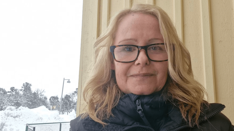 En kvinna med glasögon och mörk vinterjacka står utomhus framför en gul trävägg. I bakgrunden syns snötäckt mark, träd och en gatlykta.