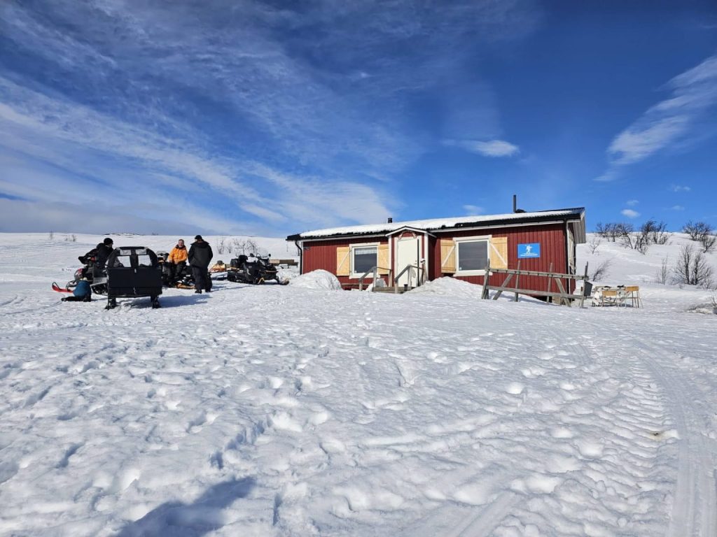 Snötäckt landskap men en röd trästuga i förgrunden och personer med snöskotrar utanför.