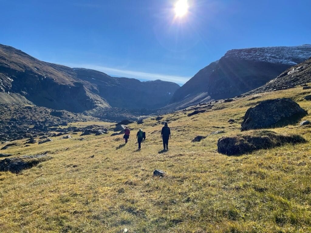 Tre vandrare går genom en gräsbevuxen fjälldal omgiven av branta berg, med solen högt på en klarblå himmel.