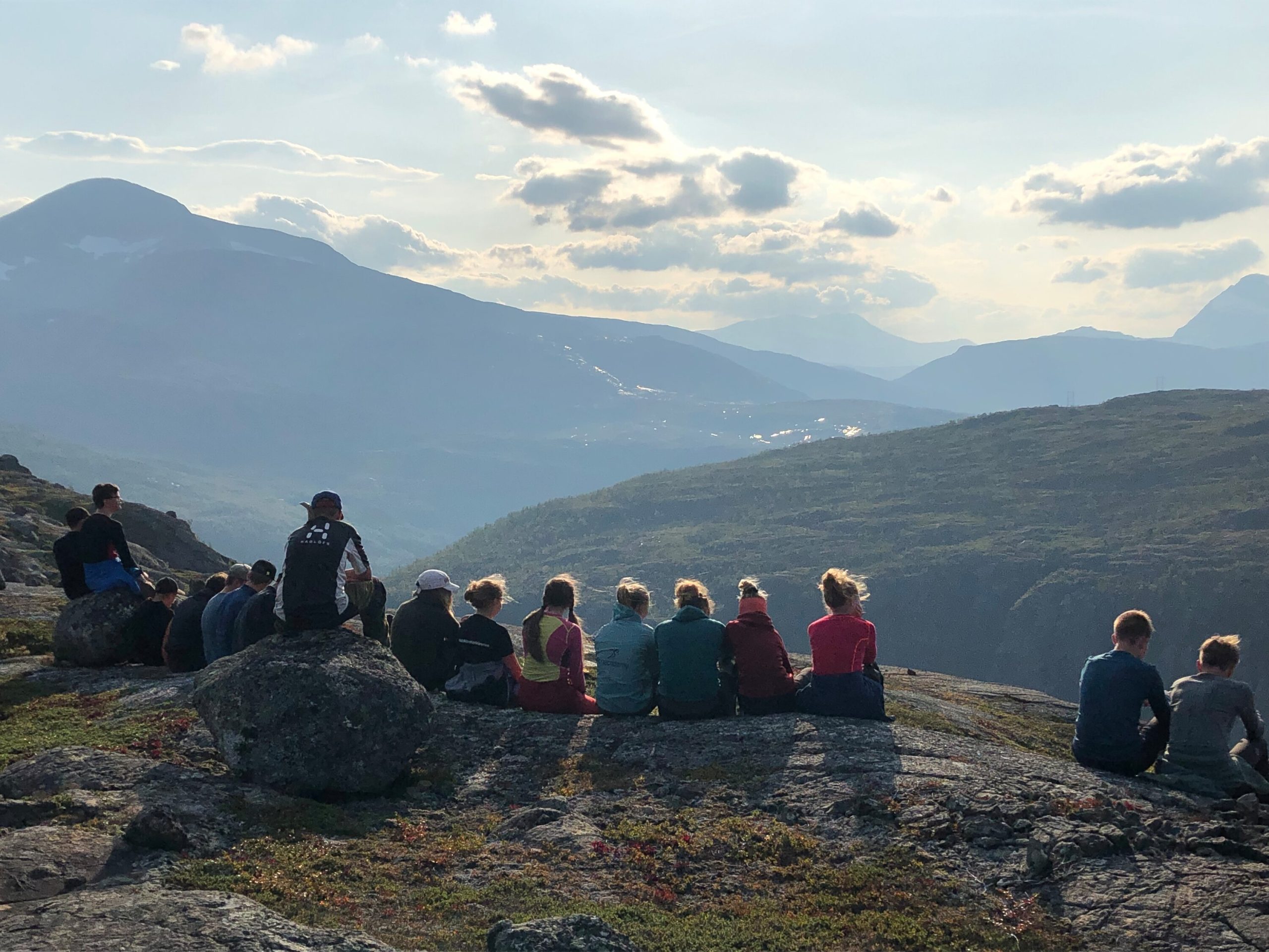 En grupp ungdomar sitter samlade på klipphällar och blickar ut över ett vidsträckt fjällandskap i kvällsljus, troligen under en fjällvandring.