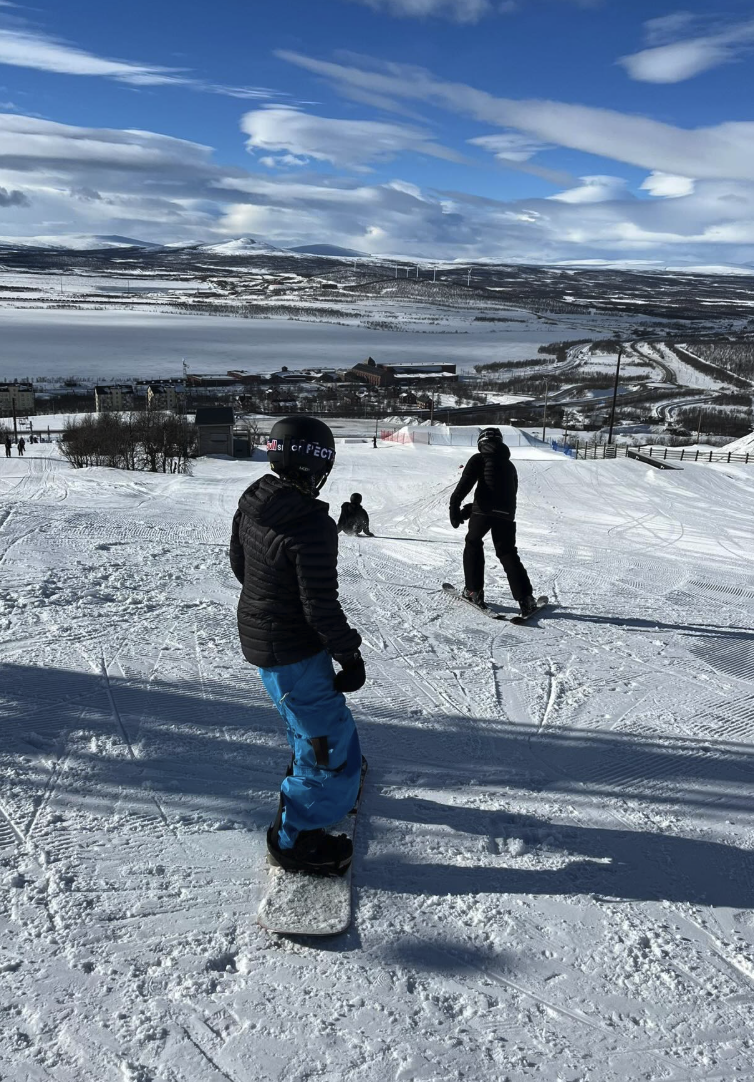 Vy uppifrån en skidbacke med en person på snowboard i förgrunden, klädd i svart jacka, blå byxor och svart hjälm. Längre ner i backen står en person på skidor. I bakgrunden syns en vidsträckt vinterlandskap med en liten stad, en sjö, kullar och ett soligt, delvis molnigt himmel med vindkraftverk vid horisonten.